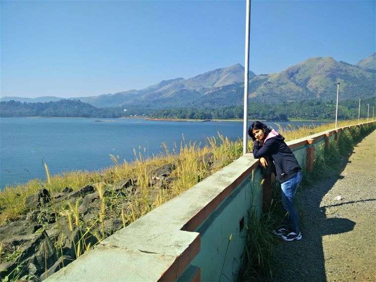 Me in a navy blue jacket at the roadside overlooking one of the most beautiful lakes in India with green mountains in the backdrop